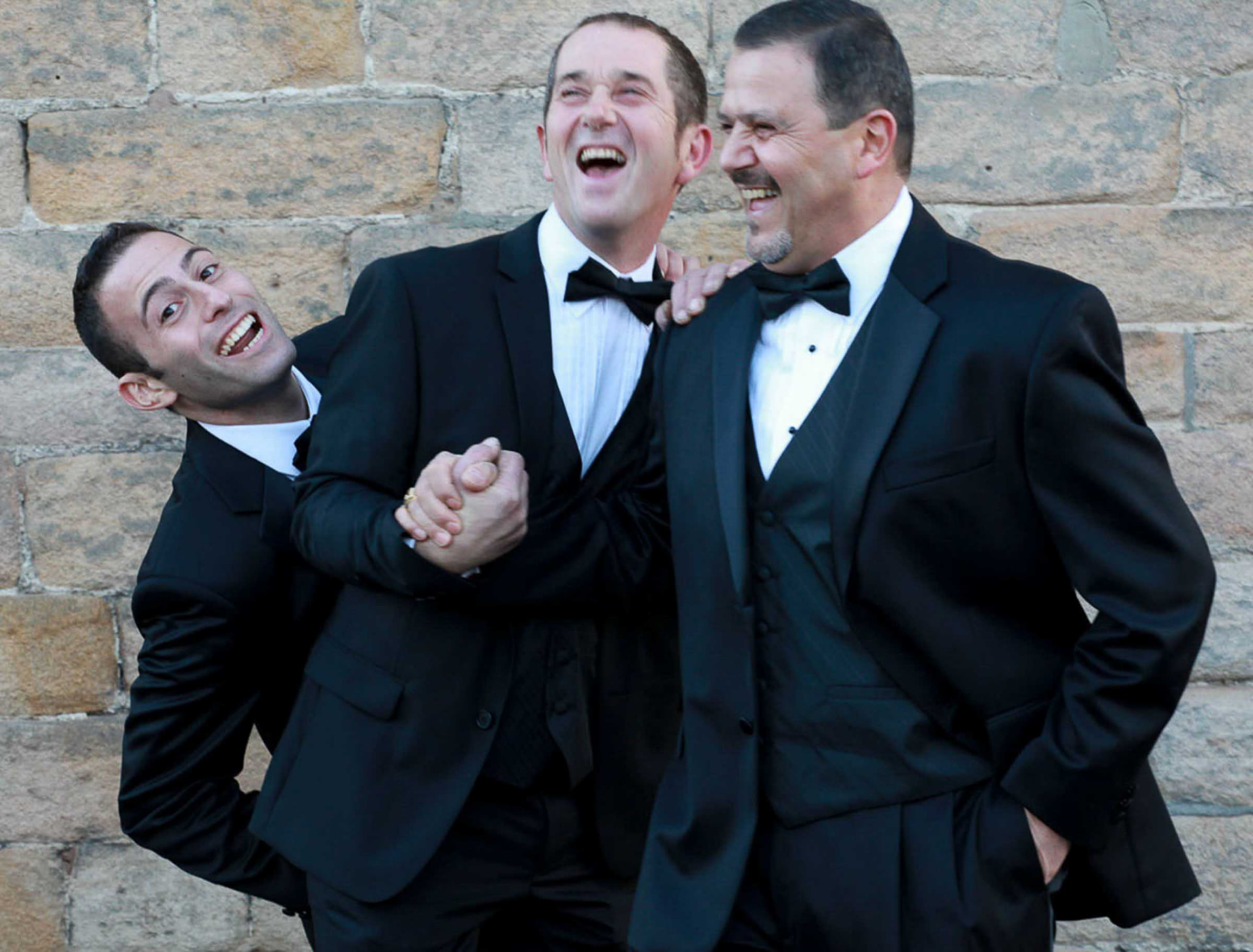 Three groomsmen in black tie laughing against a stone wall at a Newport wedding