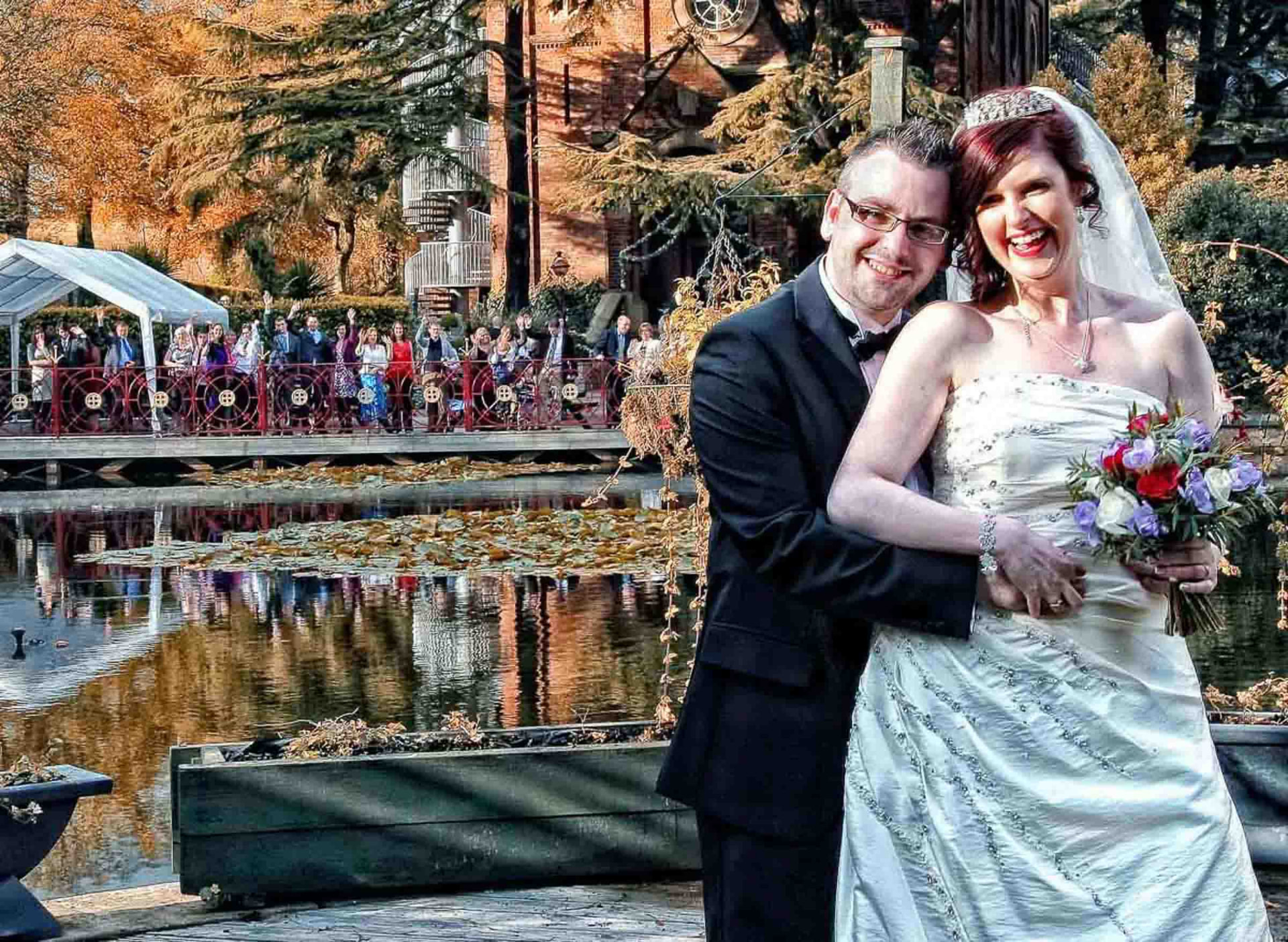 Bride and groom laughing by a lake as guests cheer from a bridge, South Wales wedding photographer